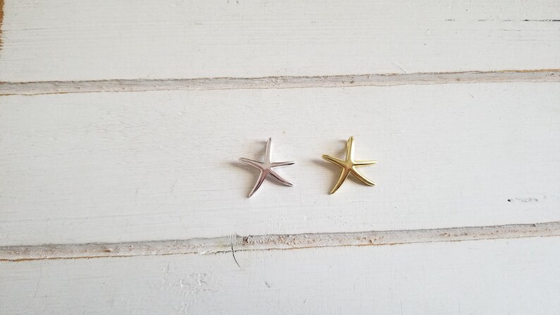 May include: Two starfish charms, one silver and one gold, on a white wooden background.