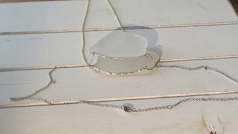 Op de afbeelding: Een delicate ketting met een gouden ketting en een horizontale hanger versierd met kleine, sprankelende steentjes. Een tweede, zilveren ketting is ook zichtbaar, waardoor een gelaagd effect ontstaat. De sieraden worden op een wit oppervlak gepresenteerd.