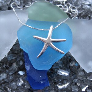 May include: A silver starfish pendant necklace on a silver chain. The necklace is displayed on a bed of blue, green, and white sea glass.