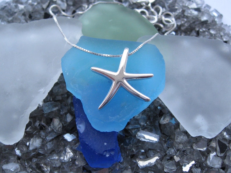 May include: A silver starfish pendant necklace on a silver chain. The necklace is displayed on a bed of blue, green, and white sea glass.