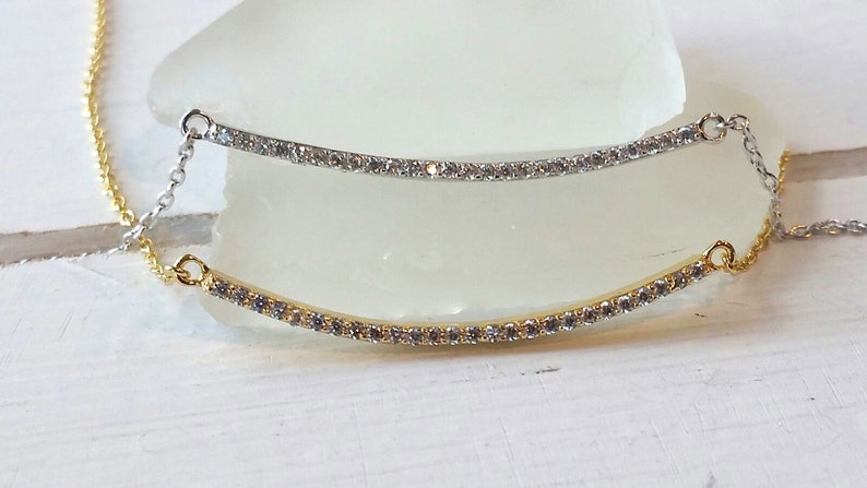 Op de afbeelding: Twee delicate kettingen met gebogen staven, &eacute;&eacute;n goudkleurig en &eacute;&eacute;n zilverkleurig, elk versierd met kleine, sprankelende stenen. De kettingen zijn verbonden door fijne kettingen, waardoor een gelaagd effect ontstaat. De sieraden worden op een wit oppervlak gepresenteerd.