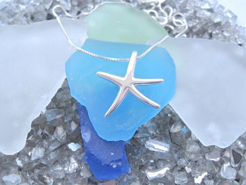 May include: A silver starfish pendant necklace on a silver chain. The necklace is displayed on a bed of blue, green, and white sea glass.