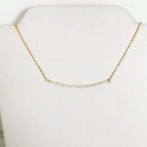 Op de afbeelding: Een goudkleurige ketting met een gebogen hanger. De hanger is bezet met kleine, heldere edelstenen. De ketting wordt getoond op een witte mannequin buste tegen een witte achtergrond. De ketting is delicaat en bestaat uit kleine schakels.