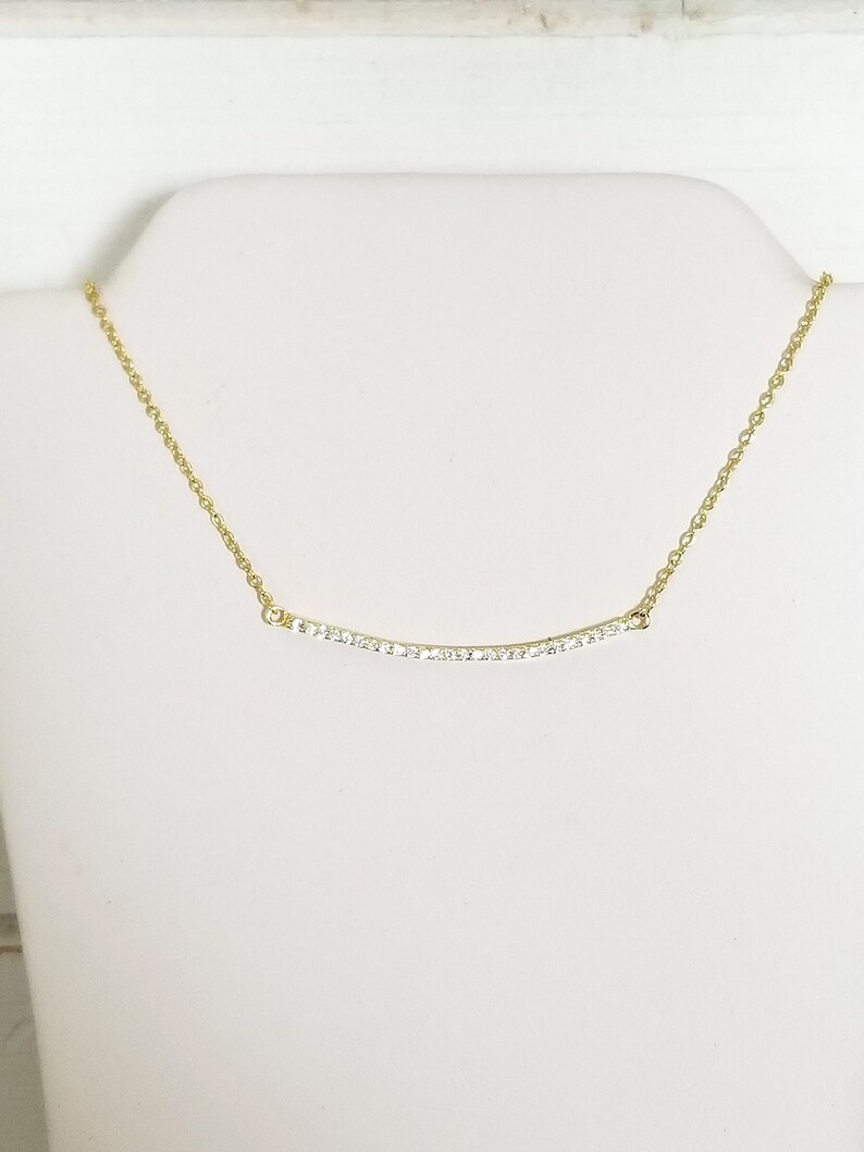 Op de afbeelding: Een goudkleurige ketting met een gebogen hanger. De hanger is bezet met kleine, heldere edelstenen. De ketting wordt getoond op een witte mannequin buste tegen een witte achtergrond. De ketting is delicaat en bestaat uit kleine schakels.