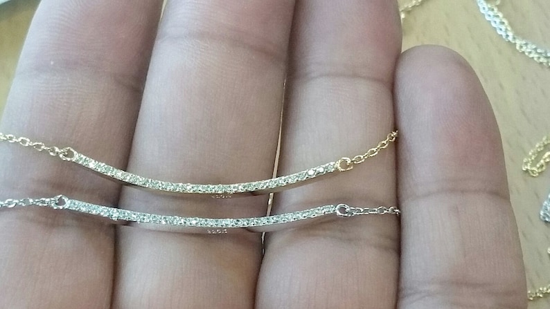 Op de afbeelding: Twee delicate gebogen stavenkettingen, &eacute;&eacute;n goudkleurig en &eacute;&eacute;n zilverkleurig, elk versierd met een rij kleine, sprankelende edelstenen. De kettingen worden gepresenteerd tegen een neutrale achtergrond, die hun elegante ontwerp benadrukt.
