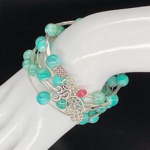 Puede incluir: Un brazalete turquesa y plateado con un dije de mano de hamsa y una cuenta rosa. El brazalete está hecho de múltiples hilos de alambre envueltos con cuentas turquesas.