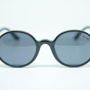 May include: Oval sunglasses in black with dark grey lenses and gold-coloured detailing on the temples. These sunglasses have a classic design. The brand name "ROLLING" is visible on the lens. Suitable for various occasions.