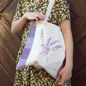 Op de afbeelding: Een vrouw in een gele en zwarte bloemenjurk houdt een witte tote bag vast met een paars lavendelontwerp. De tas heeft een paarse en witte gestreepte rand.