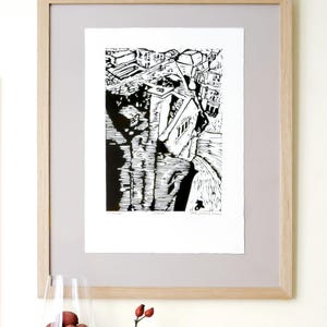The image shows a suggestion for framing the Oxford linocut print. Here, the print is placed on a grey background. It is under a glass, within a wooden frame. There is a wine gIass with litchi fruit in front of the frame. To the right, a rosehip.