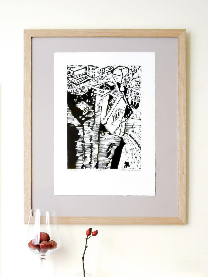 The image shows a suggestion for framing the Oxford linocut print. Here, the print is placed on a grey background. It is under a glass, within a wooden frame. There is a wine gIass with litchi fruit in front of the frame. To the right, a rosehip.