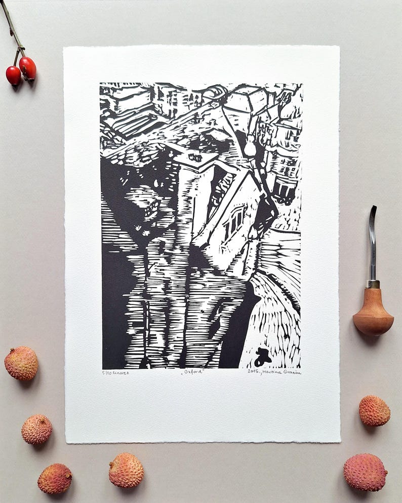 The photo shows a linocut - a birds-eye view from the Carfax tower in Oxford, England. The print is placed on a grey background. To its right is a Pfeil gouge, a tool for carving linocuts. There are some  litchi fruit around the print and a rosehip.
