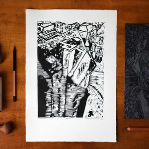 The image shows the print in a  styled scene. It is placed on a brown wooden table. To the left of it are Pfeil gouges used for carving linocuts, a pencil and a brayer. To the right of it is another pencil and the carved linocut block, the matrix.