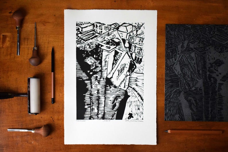 The image shows the print in a  styled scene. It is placed on a brown wooden table. To the left of it are Pfeil gouges used for carving linocuts, a pencil and a brayer. To the right of it is another pencil and the carved linocut block, the matrix.