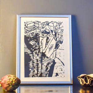 The image shows a suggestion for framing the print. Here it is framed in a silver frame and shot against a grey wall. In front of it, to the right is an artichoke, and to the right, a  small black-and-white clay vessel. The light is coming from left.