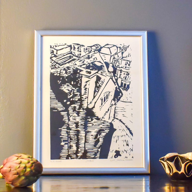 The image shows a suggestion for framing the print. Here it is framed in a silver frame and shot against a grey wall. In front of it, to the right is an artichoke, and to the right, a  small black-and-white clay vessel. The light is coming from left.