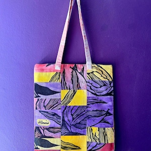 May include: A colorful tote bag with a patchwork design featuring botanical linocut prints in shades of purple, yellow, and black. The bag has long handles and a small label that reads "H. Gracie". The background is a solid purple.