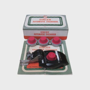 May include: A black Singer Automatic Zigzagger sewing machine attachment with a red dial, three additional red and silver circular cams, and a rectangular box labeled SINGER AUTOMATIC ZIGZAGGER. The set includes a printed instruction manual.