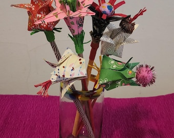 May include: A clear glass vase holds a collection of colorful origami flowers. The flowers are made from patterned paper in shades of red, pink, green, and cream. Each flower has a unique design and is attached to a wooden stem.
