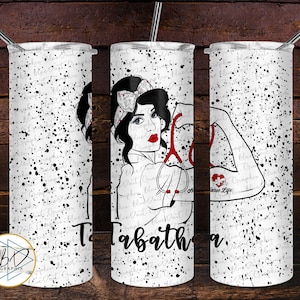 May include: A white tumbler with a black splatter pattern and a design of a woman with a stethoscope and the text "Nurse Life" and "Tabatha".