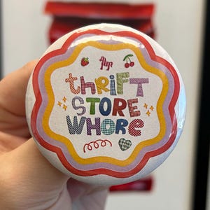 Könnte beinhalten: Ein runder Button mit dem Text "thrift store whore" in bunten Buchstaben. Der Button hat einen weißen Hintergrund mit einem gewellten Rand und einem Regenbogenfarbenen Rahmen. Verzierungen sind Kirschen und Sterne.