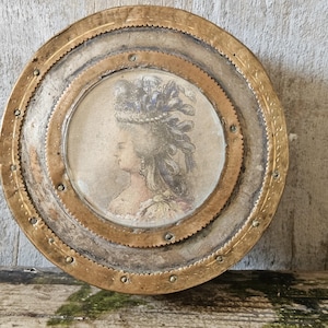 May include: Vintage circular decorative plate featuring a profile portrait of a woman. The plate has a brass and silver-toned metal frame with a decorative edge. The portrait is centrally positioned.