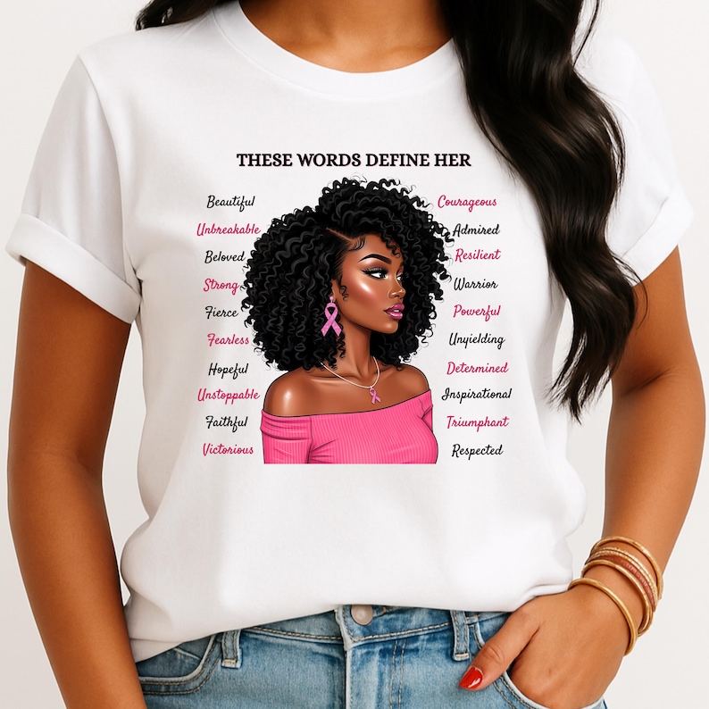 May include: White t-shirt featuring a graphic of a woman with curly black hair, wearing a pink off-the-shoulder top and a pink ribbon. The text "THESE WORDS DEFINE HER" and a list of empowering words are printed on the shirt.