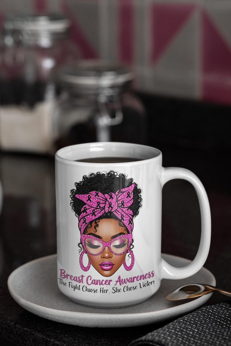 May include: White ceramic mug featuring a woman with a pink headscarf and glasses, designed for breast cancer awareness. The mug includes the text "Breast Cancer Awareness The Fight Chose Her. She Chose Victory!" on the front. The mug is on a white saucer.