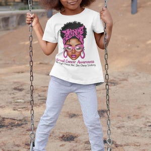May include: A young girl wearing a white t-shirt with a graphic supporting Breast Cancer Awareness. The graphic features a woman with pink glasses and a pink headscarf. The text reads "Breast Cancer Awareness The Fight Chose Her, She Chose Victory."