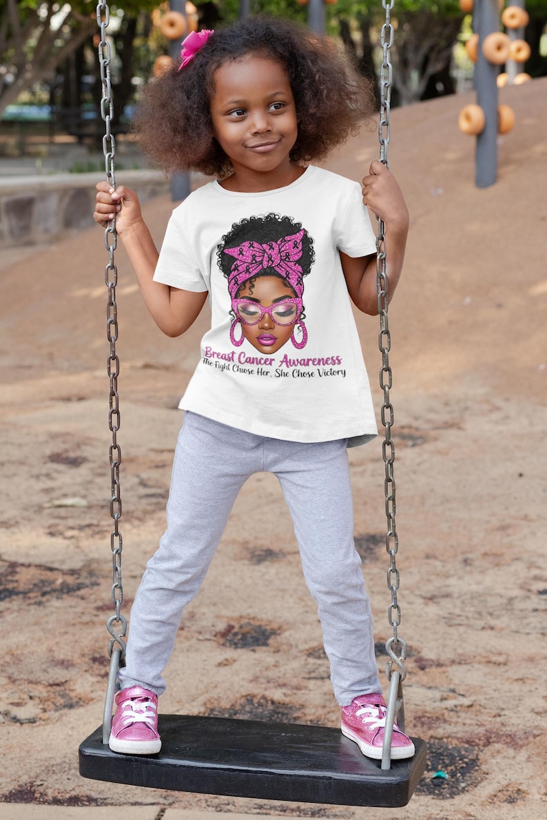 May include: A young girl wearing a white t-shirt with a graphic supporting Breast Cancer Awareness. The graphic features a woman with pink glasses and a pink headscarf. The text reads "Breast Cancer Awareness The Fight Chose Her, She Chose Victory."