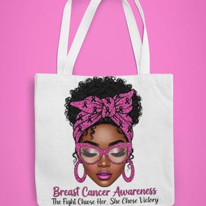 May include: White tote bag featuring a graphic of a woman with a pink headscarf and glasses, adorned with pink ribbons. The text "Breast Cancer Awareness" and "The Fight Chose Her, She Chose Victory" are below the image. The bag has white handles.