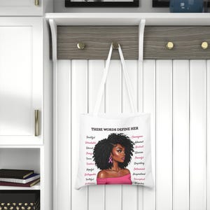 May include: White tote bag featuring a portrait of a woman with curly black hair and pink accents. The text "THESE WORDS DEFINE HER" is above the portrait, with a list of empowering words in pink on the sides.