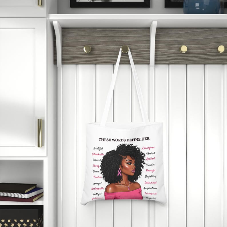 May include: White tote bag featuring a portrait of a woman with curly black hair and pink accents. The text "THESE WORDS DEFINE HER" is above the portrait, with a list of empowering words in pink on the sides.