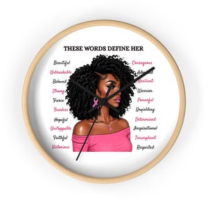 May include: A round wall clock with a light wood frame. The clock face features an illustration of a woman with curly black hair and pink accents. Text around the image reads "THESE WORDS DEFINE HER" and lists empowering words.