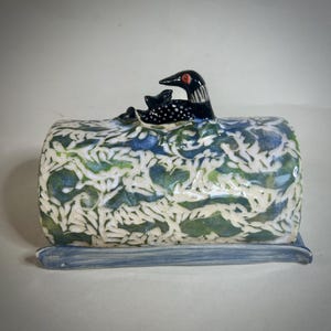 May include: A ceramic butter dish with a cylindrical cover featuring a glossy blue and green glaze with white leaf-like patterns. A sculpted black and white loon with a red eye sits on top of the cover, resting on a flat blue rectangular base.