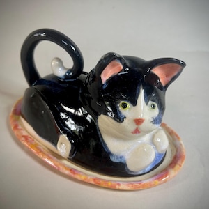 May include: A ceramic butter dish shaped like a black and white cat lying down. The cat has a curled tail, pink-lined ears, and yellow eyes. It rests on an oval-shaped base with a mottled orange and yellow rim, featuring a glossy finish.