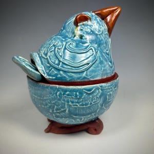 May include: A ceramic bird-shaped container with a lid. The container is light blue with embossed patterns and a brown beak and base. The lid is slightly ajar, revealing the interior.