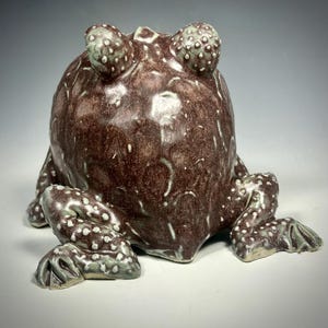 Puede incluir: Una escultura de cer&aacute;mica de rana con un esmalte marr&oacute;n y verde. La rana est&aacute; sentada sobre sus patas traseras con las patas delanteras metidas. La rana tiene una textura abultada y ojos grandes.