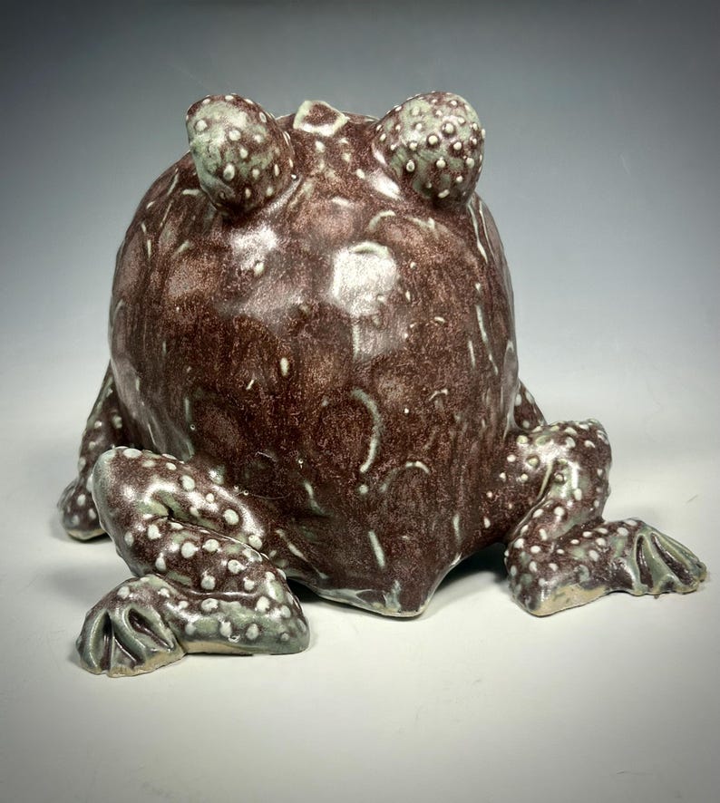 Puede incluir: Una escultura de cer&aacute;mica de rana con un esmalte marr&oacute;n y verde. La rana est&aacute; sentada sobre sus patas traseras con las patas delanteras metidas. La rana tiene una textura abultada y ojos grandes.