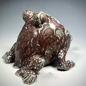 Puede incluir: Una escultura de cer&aacute;mica de rana con un esmalte moteado marr&oacute;n y blanco. La rana est&aacute; sentada sobre sus patas traseras con las patas delanteras extendidas.