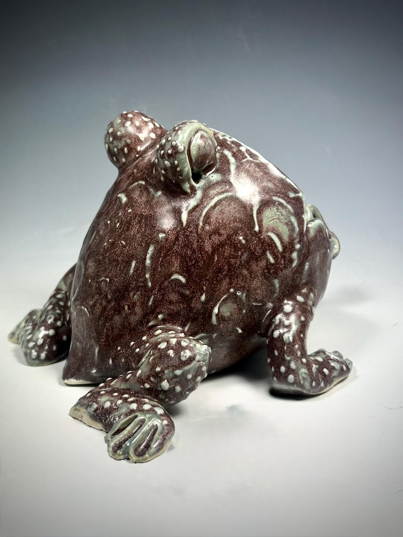 Puede incluir: Una escultura de cer&aacute;mica de rana con un esmalte moteado marr&oacute;n y blanco. La rana est&aacute; sentada sobre sus patas traseras con las patas delanteras extendidas.