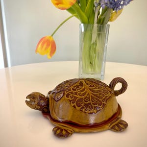 May include: A ceramic turtle-shaped trinket box with a detailed, leaf-like pattern on its shell. The box is primarily tan with dark brown accents and a red rim. The turtle has a curled tail and is on a white surface.