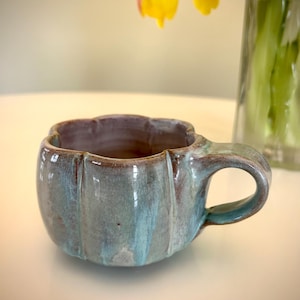 May include: A handmade ceramic mug with a unique, textured design. The mug features a teal and brown glaze, with a rounded shape and a handle. The interior is a darker shade of brown. A yellow and orange flower is in the background.