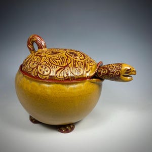 May include: A ceramic turtle-shaped container with a lid. The container is primarily yellow with brown and red accents. The lid features an intricate carved design. The turtle's head extends from the container, and it has small feet.