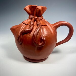 May include: A terracotta-colored teapot with a unique design. The teapot is shaped like a tied bag, with a handle and spout. The lid is also designed to look like the top of the bag, with a tied knot. The teapot is made of ceramic.