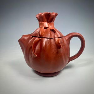 May include: A reddish-brown ceramic teapot with a unique design. The teapot is shaped like a bag tied at the top with a ribbon. It has a curved handle and a spout for pouring. The teapot sits on a small, round base.