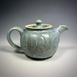 May include: A small, light blue ceramic teapot with a textured, leaf-like design. The teapot has a lid and a handle.