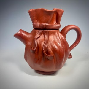 May include: A reddish-brown ceramic teapot with a unique design. The teapot has a rounded body with a spout, handle, and a lid that resembles a tied bag. The overall shape is organic and textured, suggesting a handmade quality. The teapot is likely used for brewing tea.