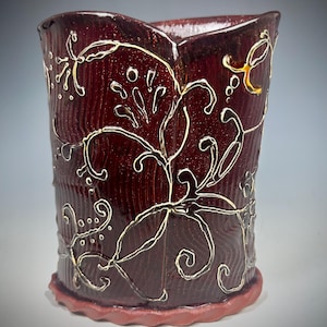May include: A burgundy ceramic vase with a textured surface and a scalloped rim. The vase is adorned with a white, swirling floral design. The base of the vase is a darker shade of burgundy.