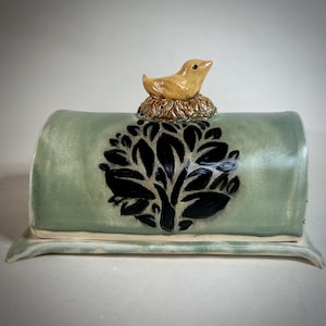 May include: A ceramic butter dish with a light green glazed finish and a dark black tree silhouette design. The lid features a sculpted, tan-colored bird resting in a textured nest, serving as a handle for the rectangular, curved cover.