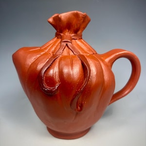 May include: A terracotta-colored teapot with a unique design. The teapot features a handle, spout, and a lid shaped like a tied bag. The surface has textured details and a rich, earthy tone, suggesting a handmade ceramic piece.